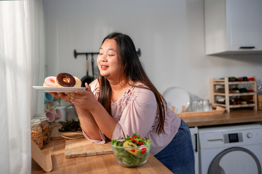 Asian plus-size woman reject salad and chooses to eat donut instead.  - Powered by Adobe