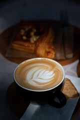 A cup of hot latte art coffee served with biscuit and pastry on wooden table in cozy cafe atmosphere with natural morning light.