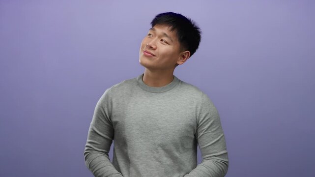 Young man in gray sweater stands against purple background with a serene expression, eyes closed and slight smile, conveying peaceful and relaxed mood.
