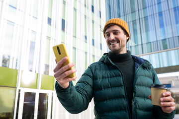 Smiling man using smartphone and holding coffee outside office building