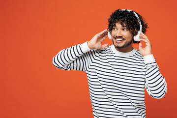 Young smiling happy Indian man he wears striped sweatshirt casual clothes listen to music in headphones look aside on area isolated on plain red orange background studio portrait. Lifestyle concept.