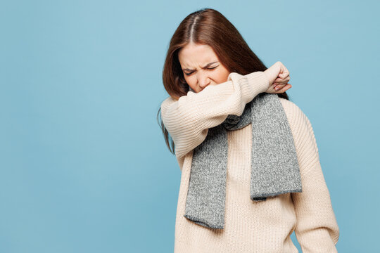 Young woman wears pink sweater scarf sneeze into her elbow isolated on plain pastel light blue background studio portrait Healthy lifestyle ill sick disease treatment recuperation cold season concept