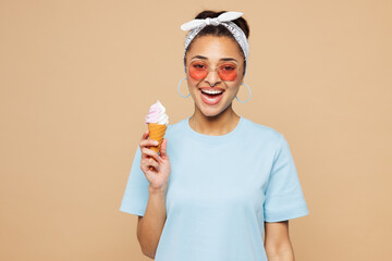 Young smiling happy woman of African American ethnicity wear blue t-shirt casual clothes hold in hand eat vanilla ice-cream isolated on plain pastel light beige background studio. Lifestyle concept.