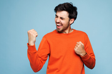 Young excited overjoyed man wear orange sweatshirt casual clothes doing winner gesture celebrate clenching fists say yes isolated on plain pastel light blue cyan background studio. Lifestyle concept.