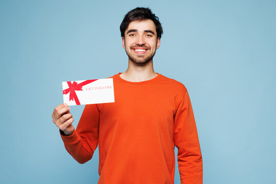 Fototapeta Young smiling happy man wear orange sweatshirt casual clothes hold gift certificate coupon voucher card for store isolated on plain pastel light blue cyan background studio portrait Lifestyle concept