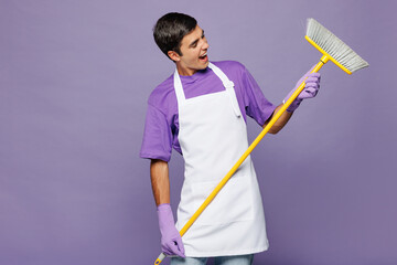 Young shocked surprised man he wears violet t-shirt hold broom mop sweeping floor while doing housework tidy up isolated on plain pastel light purple background studio portrait. Housekeeping concept.