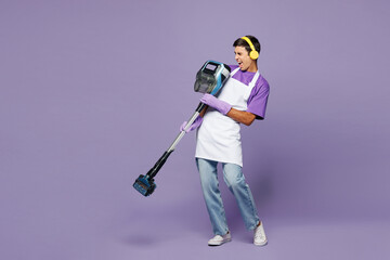 Full body young man wear violet t-shirt headphones listen music cleaning room using modern vacuum cleaner while doing housework tidy up isolated on plain pastel purple background Housekeeping concept
