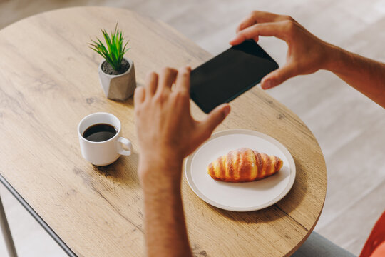 Close up cropped shot young man taking cell phone picture of croissant order sitting alone at table in coffee shop cafe relax rest in restaurant during free time indoors. Lifestyle blogging concept.