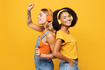Side view two young friends buddies women wear denim casual clothes together listen music in headphones stand back to back isolated on plain yellow orange background studio portrait Lifestyle concept