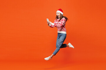 Full body side view merry young woman she wear red Christmas sweater Santa hat posing jump high use mobile cell phone isolated on plain orange background. Happy New Year celebration holiday concept.