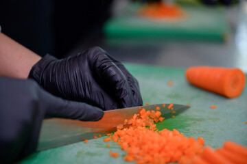 A close-up of chef hands in black gloves finely dicing carrot on green cutting board, with selective focus and soft lighting in commercial kitchen.