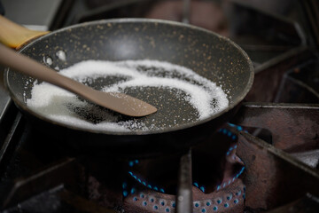 Close-up shot of sugar melting in nonstick pan over blue gas flame, concept of dessert preparation and caramel making in professional kitchen.