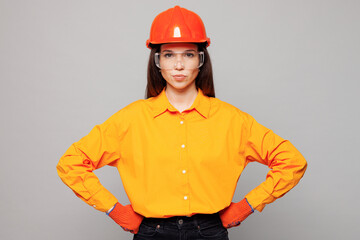 Young architect engineer designer employee woman wear yellow clothes hardhat glasses hold hands on waist stand akimbo isolated on plain grey background studio portrait. People work on build project.