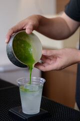 Barista pouring matcha tea into glass of soda with ice, creating refreshing layered drink in modern cafe.