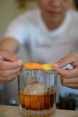 Close-up shot of bartender hands twisting orange peel over glass of cocktail with ice sphere, captured with shallow depth of field and warm soft light for premium drink concept.