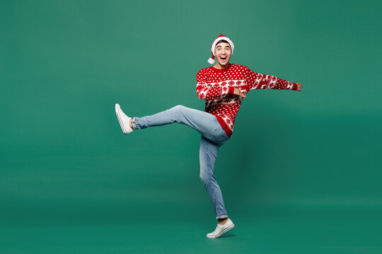 Full body side view merry young man wear red warm cozy Christmas sweater Santa Claus hat posing walk go hurry up raise up hand leg isolated on plain green background. Happy New Year holiday concept. - Powered by Adobe