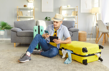Handsome cheerful young man sitting on floor with packed suitcase holding mobile phone, booking...