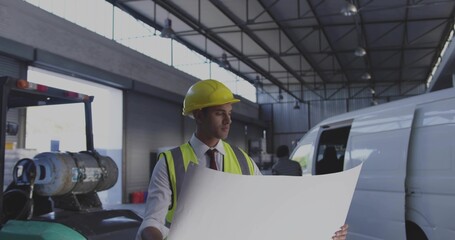 Engineer wearing safety vest and hard hat examining large blueprints at workshop, forklift, van