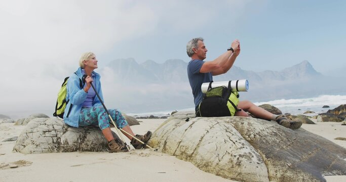 Sitting senior hiking couple capturing misty mountain view on coastal rocks, with backpacks
