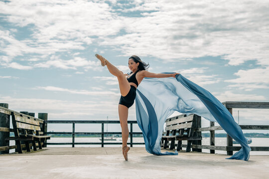 Ballet dancer lifts leg on pier with trailing sheer blue fabric