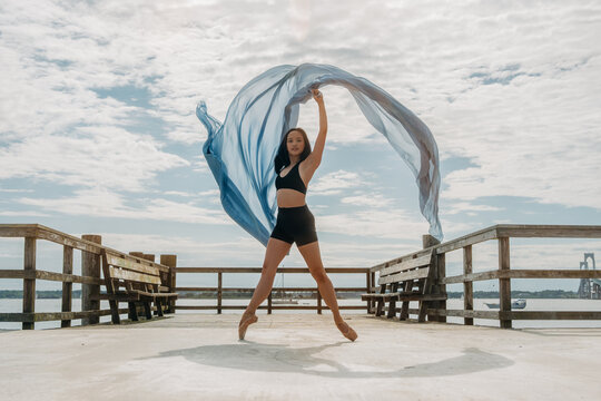 Ballerina dancing on pier holding flowing fabric against sky
