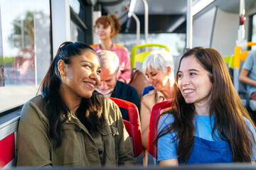 Young diverse women smiling and talking while traveling on a public bus among other passengers. Urban commuting and friendship concept