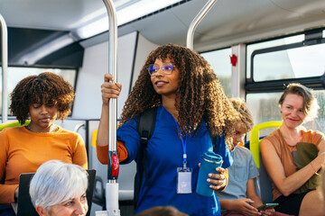 African female nurse wearing blue scrubs holding a water bottle and handrail while commuting on a public bus