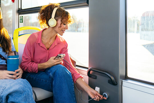Woman listening to music with headphones and charging her smartphone on a modern public bus, connecting to onboard usb