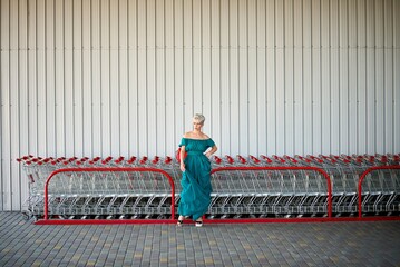 Wide shot of a woman in a teal off-shoulder dress standing by rows of red-handled shopping carts under an industrial canopy. Strong color contrast and graphic repetition.