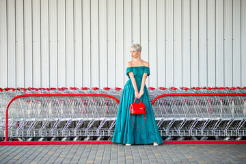 Full-length fashion shot with a bright red handbag adding a pop of color. Woman in teal off-shoulder dress stands in front of shopping carts; modern urban retail scene.