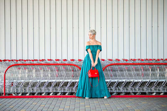 Full-length fashion shot with a bright red handbag adding a pop of color. Woman in teal off-shoulder dress stands in front of shopping carts; modern urban retail scene. - Powered by Adobe