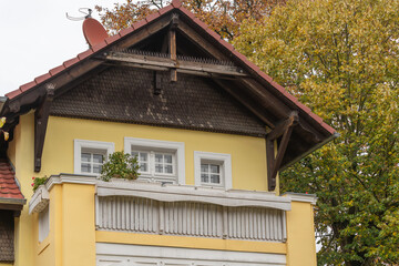 Charming yellow house with balcony and wooden roof details. Surrounded by green and orange autumn trees.