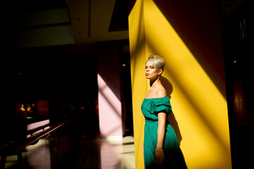 Side view portrait of woman in emerald green dress leaning on yellow wall, strong sunlight and dramatic shadows in modern interior.