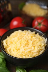 Grated cheese in bowl, tomatoes and basil on table, closeup