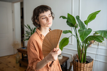 Obraz premium Woman at home suffering from summer heat, standing beside window in living room, waving hand paper fan for cooling down. Tired young female with sick expression, overwhelmed by summer heat or fever.