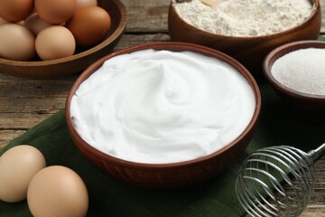 Whipped egg whites in bowl, whisk and ingredients on wooden table, closeup
