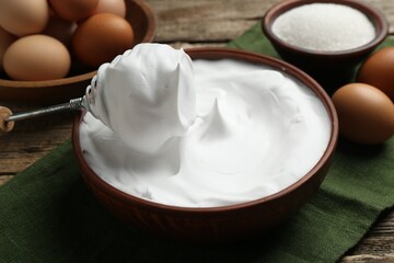Whipped egg whites in bowl, whisk and ingredients on wooden table, closeup
