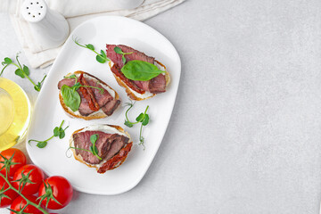 Bruschettas with roast beef, cream cheese, basil, sun-dried tomatoes and fresh vegetables on light grey table, flat lay. Space for text