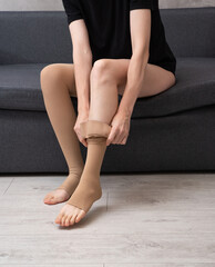 Faceless female wearing compression socks sitting on grey sofa indoors demonstrating healthy lifestyle, vein care and prevention of venous disease
