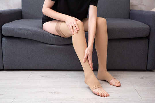 Faceless female wearing compression socks sitting on grey sofa indoors demonstrating healthy lifestyle, vein care and prevention of venous disease