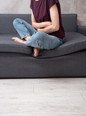 Anonymous female in casual clothes sitting indoors on grey couch holding stomach feeling abdominal pain and belly ache concept of digestive discomfort at home