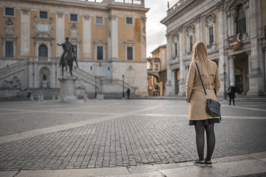 View of a solitary blonde woman in a trench coat gazes across the textured cobblestone square towards the grand architecture and equestrian statue, Rome, Lazio, Italy.