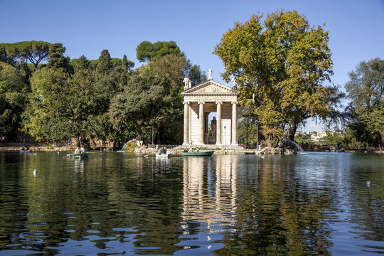 View of the Temple of Asclepius gleams in the sunlight, reflected on the still waters, framed by autumn trees, a tranquil scene, Rome, Lazio, Italy.