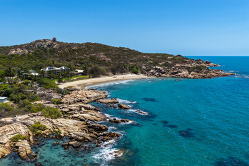 An aerial view of Rose Bay in Bowen, Queensland, reveals a stunning tropical coastline with turquoise waters, golden sand, and rocky headlands. The scene highlights coastal beauty and vibrant color.
