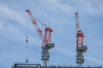 Two tower cranes dominate top of high-rise construction site. Boom extended at 45-degree angle lowers glass panel to glazing contractor on deck floor partially obscured by netted framework.