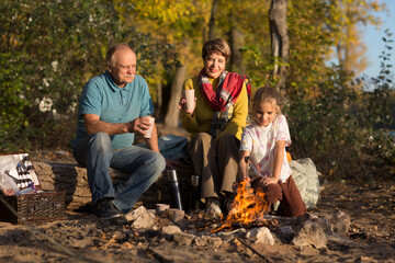 Little child girl with grandpa and grandmother Having a picnic on the beach on a sunny autumn day. Concept of friendly family.  Family  roasts marshmallows and sausages on the campfire.
