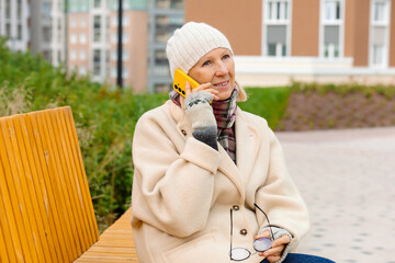 An elderly woman in warm clothing and a knitted hat sits on a wooden bench, smiling and talking on a yellow smartphone. Green bushes and residential buildings are in the background.