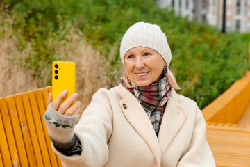 A woman wearing a light knit hat, a plaid scarf, and a beige coat sits on a yellow bench, smiling and taking a selfie on a bright yellow smartphone. City buildings are in the background.