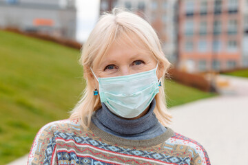A blonde woman wearing a light blue medical mask, a gray turtleneck, and a bright patterned sweater. She wears turquoise earrings. The background is a blurred cityscape with a green lawn.