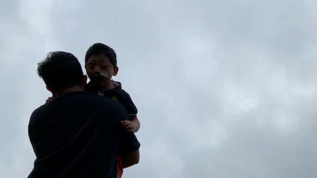 Father lifts his child, who has medical tubing, into the air against a cloudy sky, showcasing resilience and family love